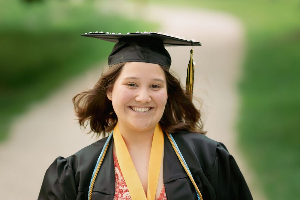 Smiling graduate in black cap and gown with yellow tassel and honor cords outdoors.