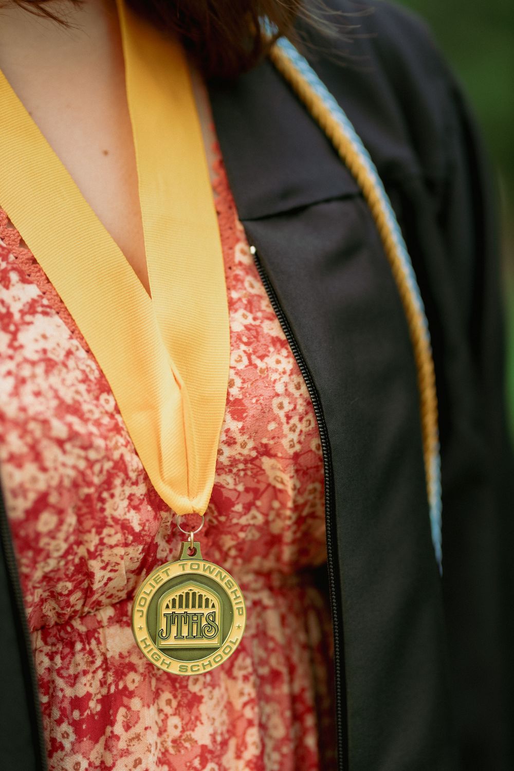 Graduate wearing gold JTHS medal on yellow ribbon over floral dress and black graduation gown.