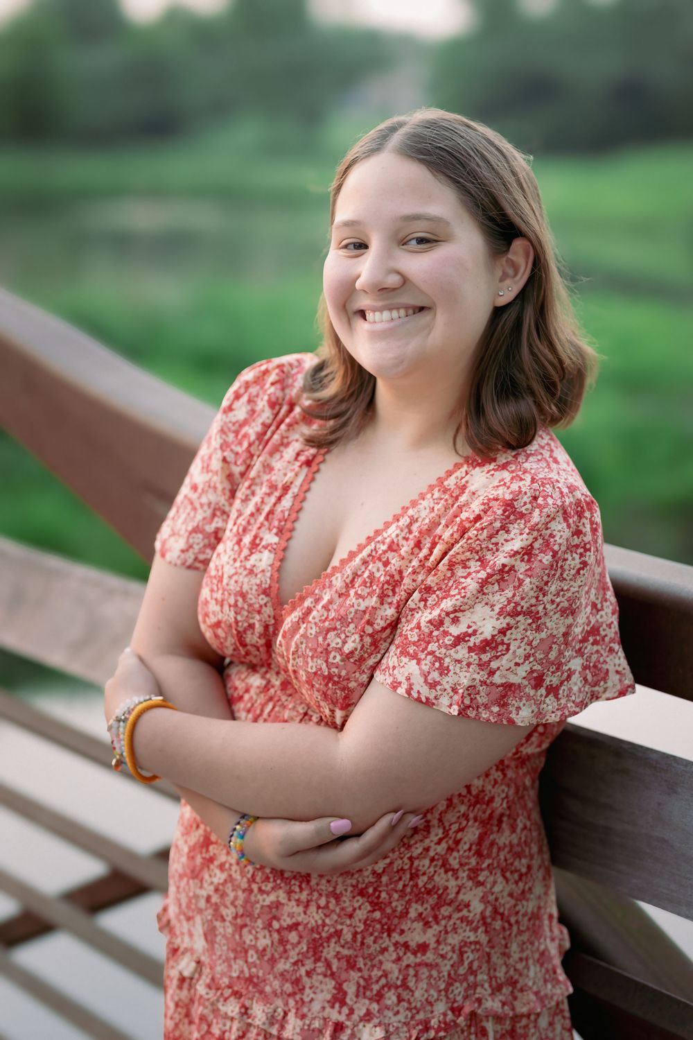 Smiling young woman in red floral dress with arms crossed leaning on wooden bridge railing.
