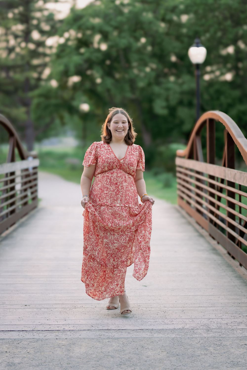 Young woman in red floral maxi dress walking on wooden bridge surrounded by lush green trees.