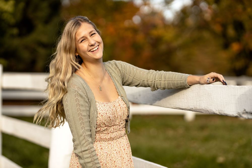 Young blonde woman in floral dress and green cardigan smiling outdoors by white fence in autumn.