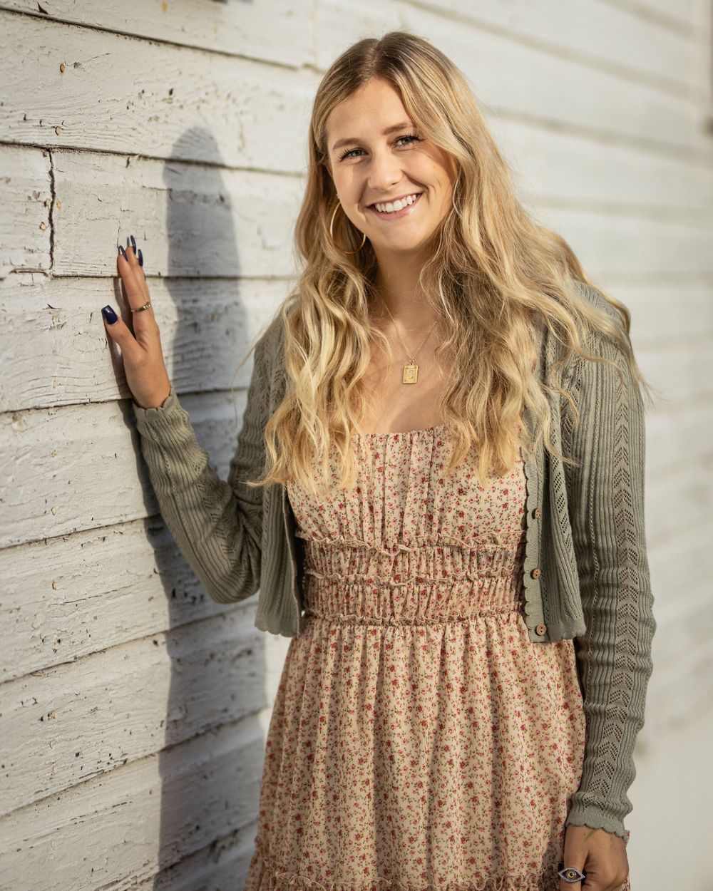 Smiling blonde woman in floral sundress and sage green cardigan leaning against white wooden wall.