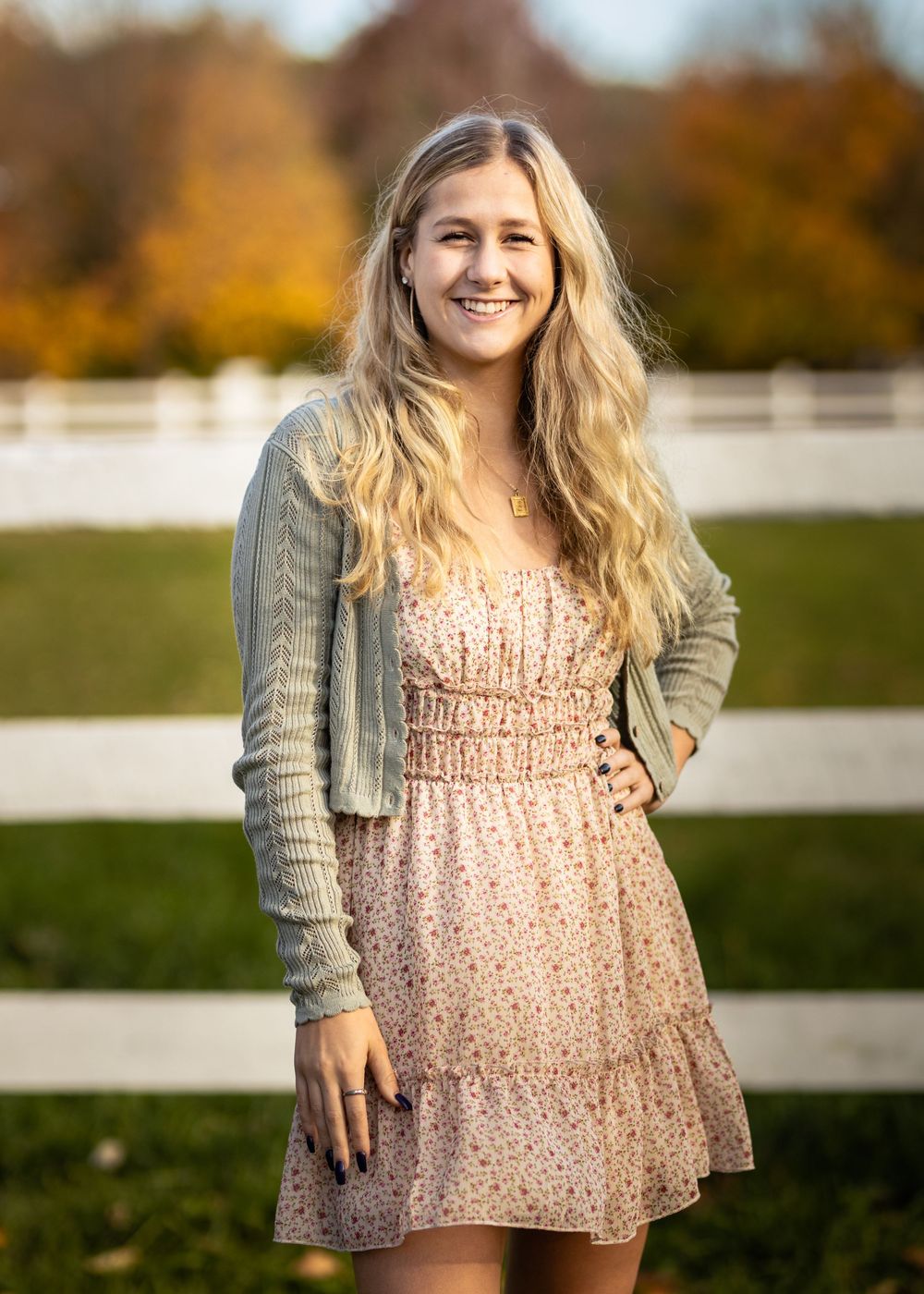 Smiling young woman in floral dress and green cardigan posing outdoors near white fence with autumn foliage.