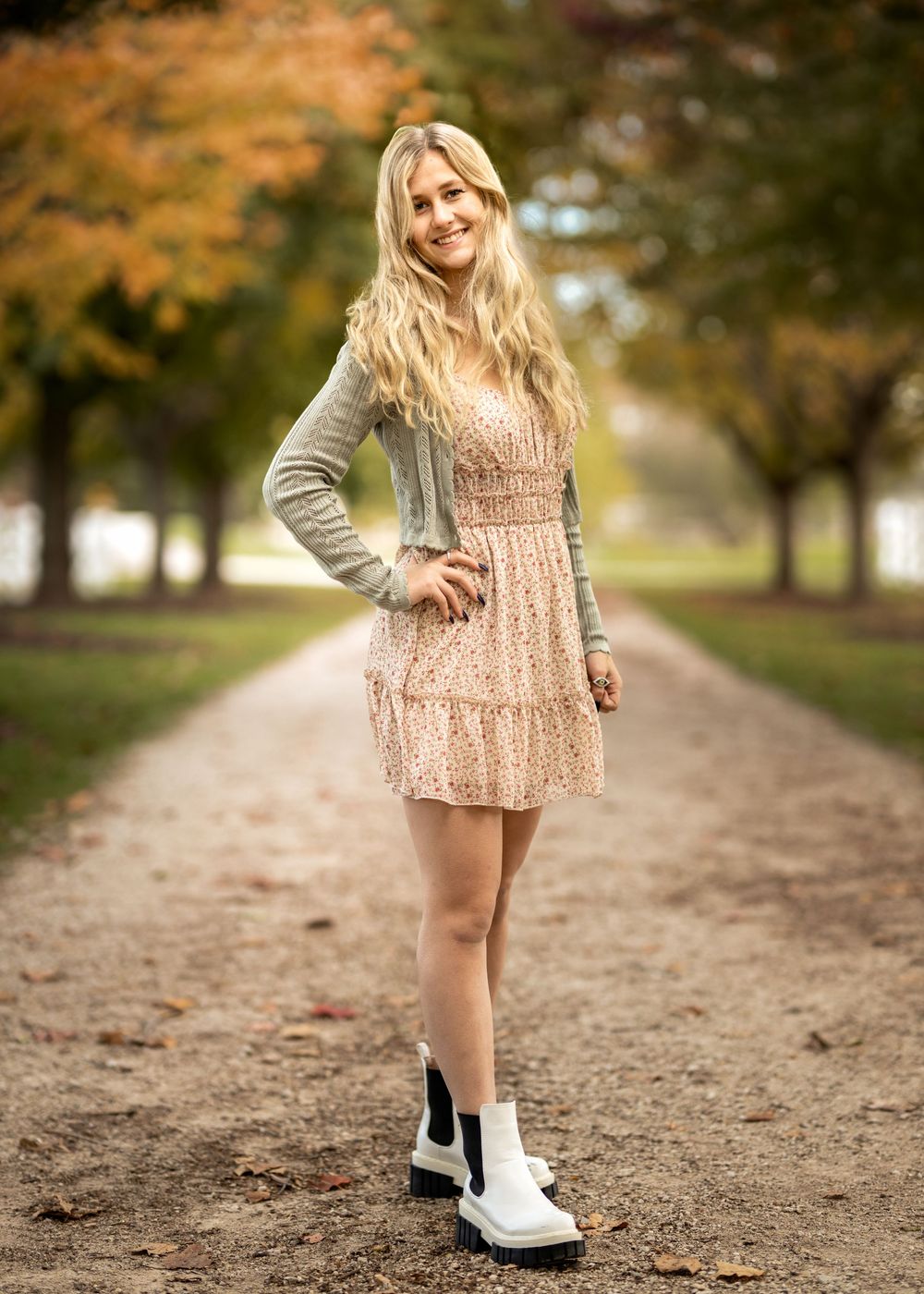 Young blonde woman in floral dress, cardigan, and white boots smiling on a tree-lined path in autumn park.