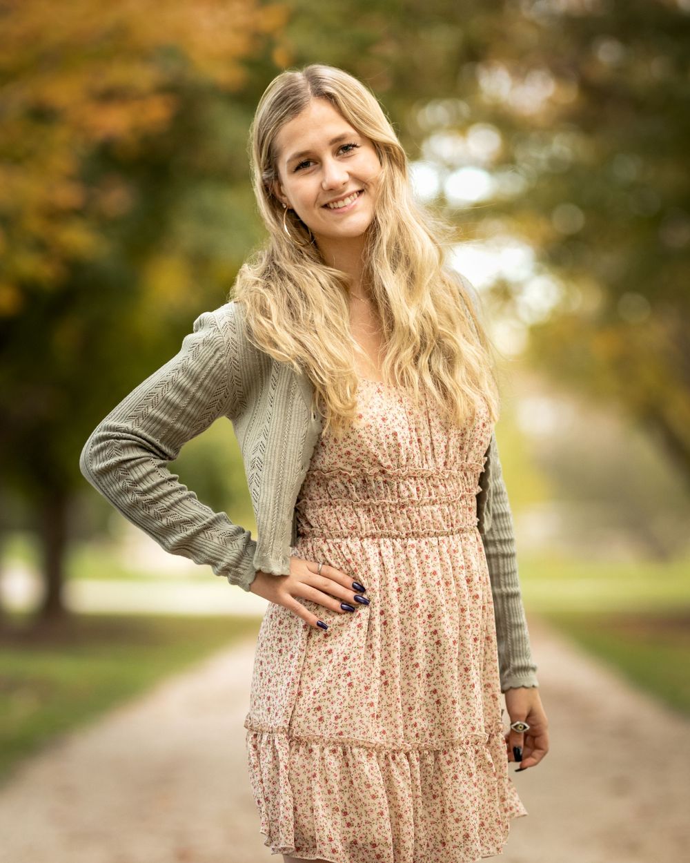 Beautiful blonde woman in floral dress and green cardigan posing on tree-lined path in autumn park.