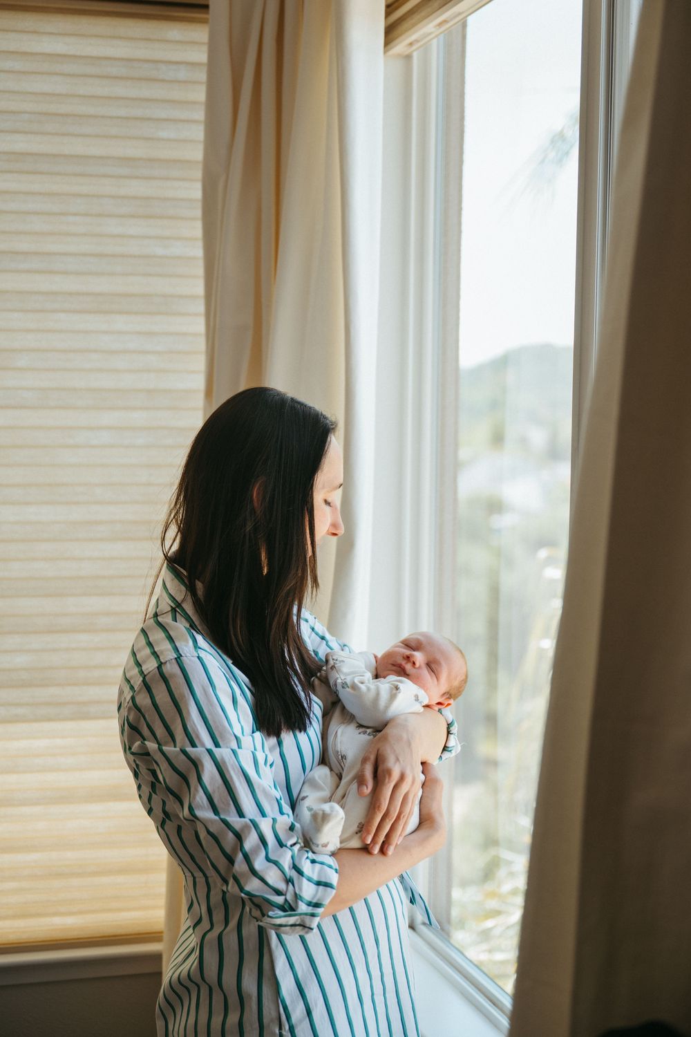 A parent in blue striped pajamas holds and gazes lovingly at their newborn baby near a sunlit window at home.