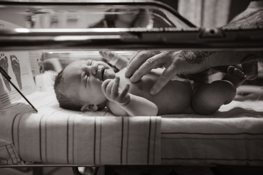 Black and white photo of a newborn baby in a hospital incubator, with an adult hand gently touching the infant.