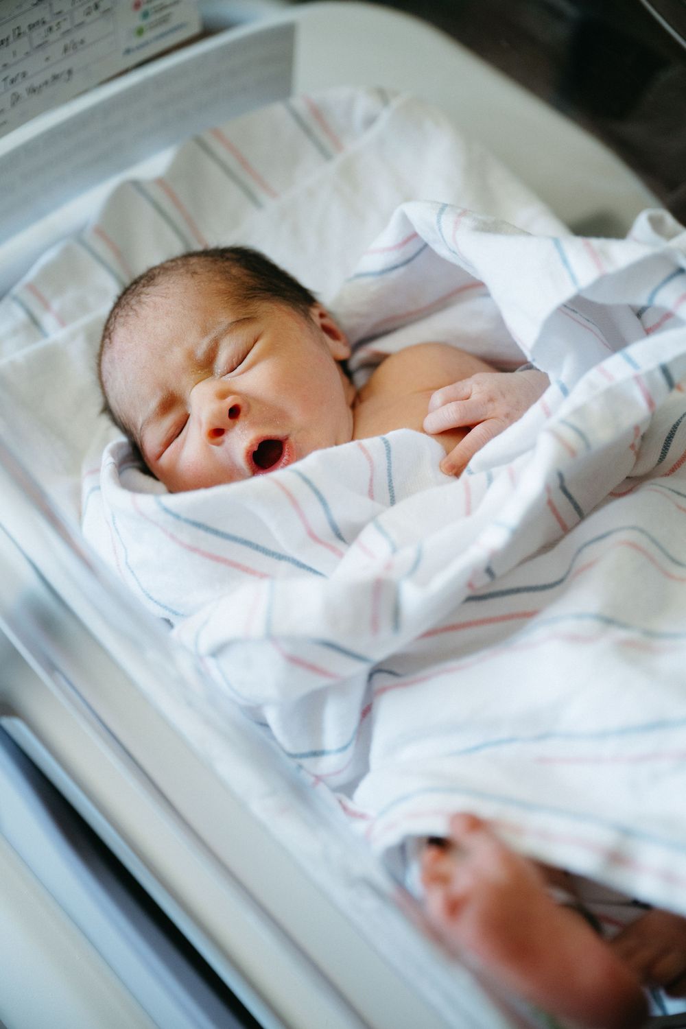 Newborn baby yawning while swaddled in striped hospital blanket lying in bassinet.