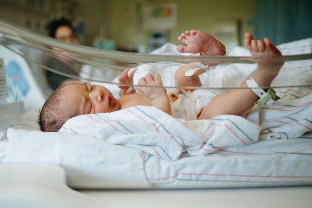 Newborn baby lying in hospital bassinet with arms raised, wrapped in striped blanket after delivery.