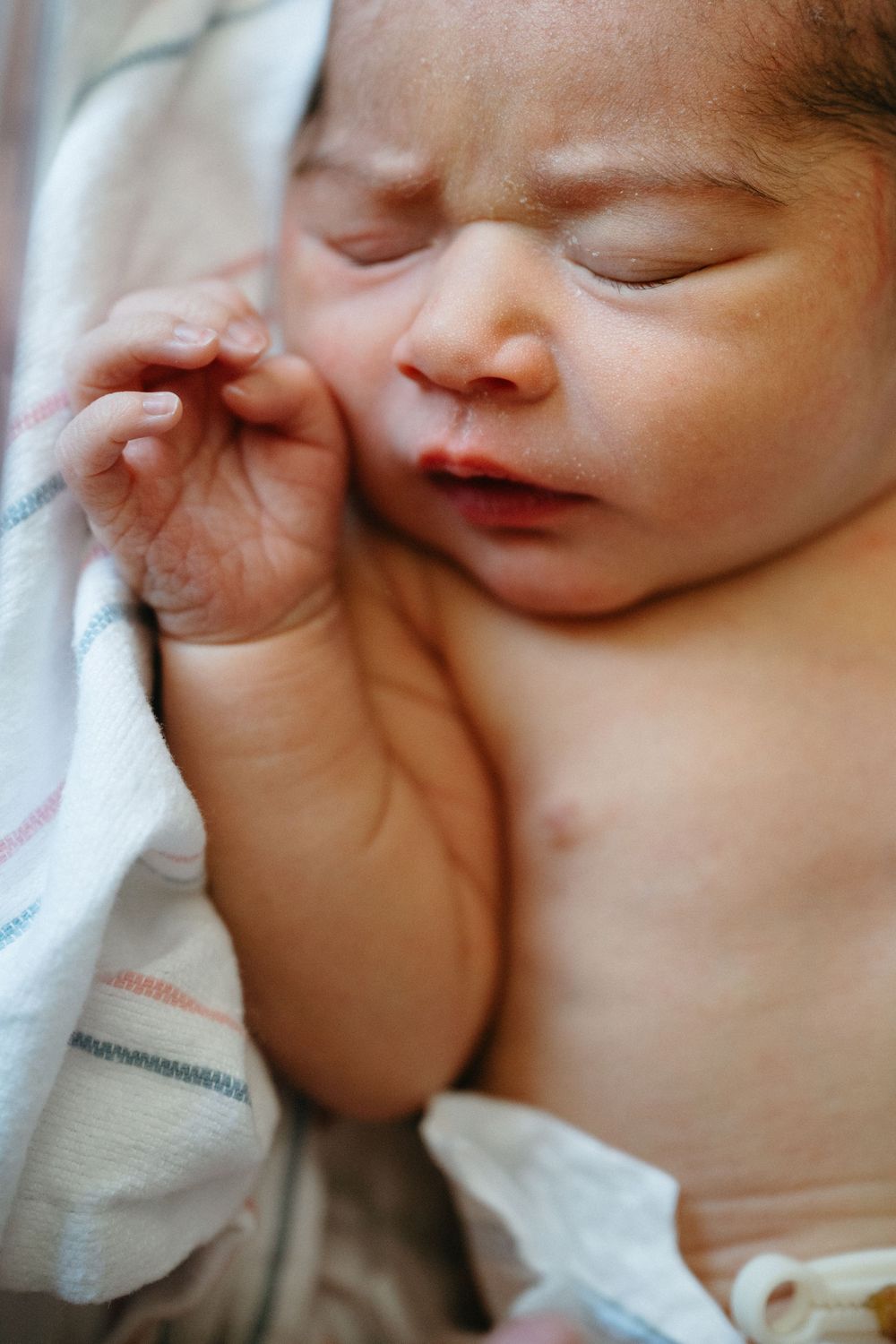 Close-up of peaceful newborn baby with tiny hand near face wrapped in white blanket.