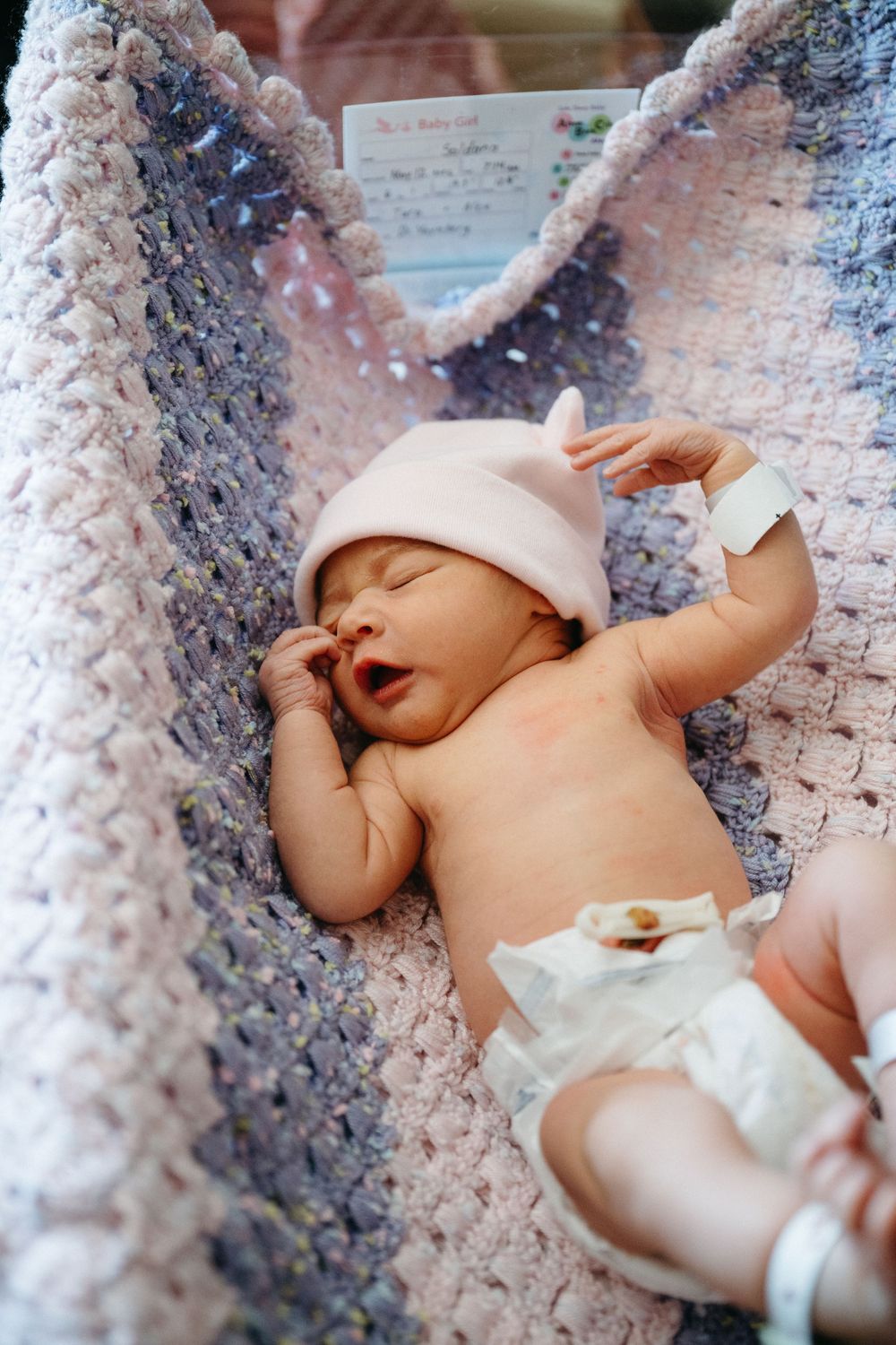 Newborn baby sleeping in a crocheted blanket wearing a pink hat and diaper in a hospital nursery.