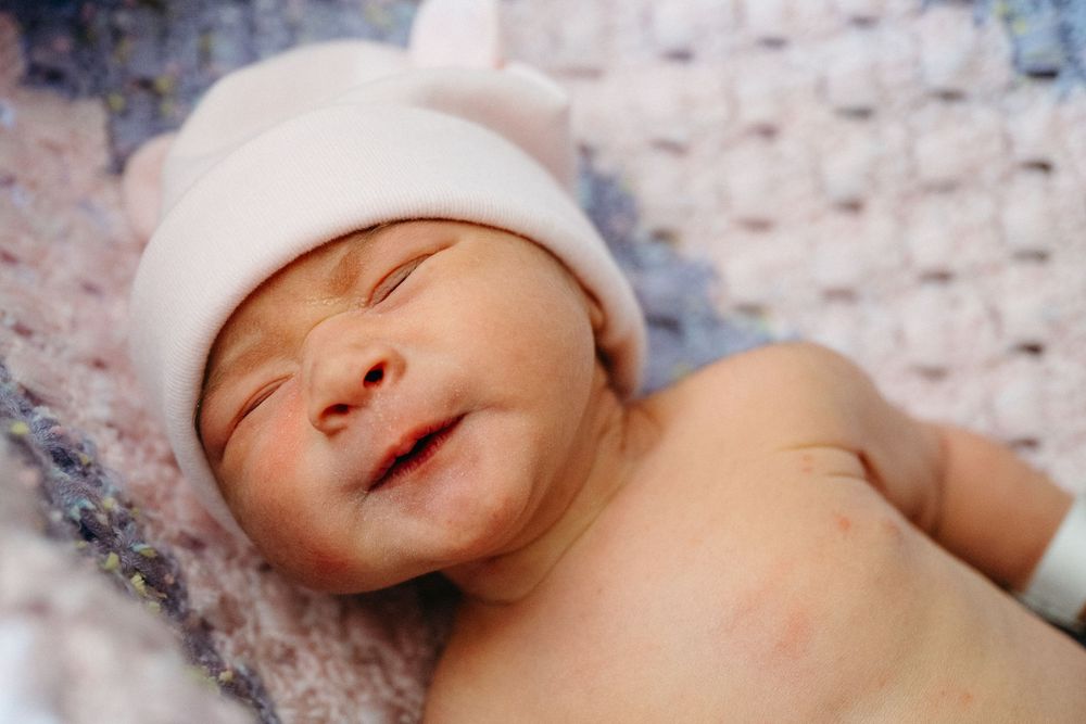 Newborn baby smiling peacefully in a white knit hat, lying on a soft blanket.