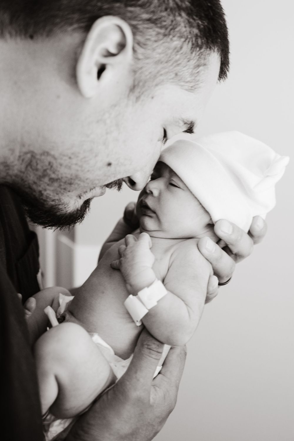 Black and white photo of a father gently kissing his newborn baby wearing a white hat.