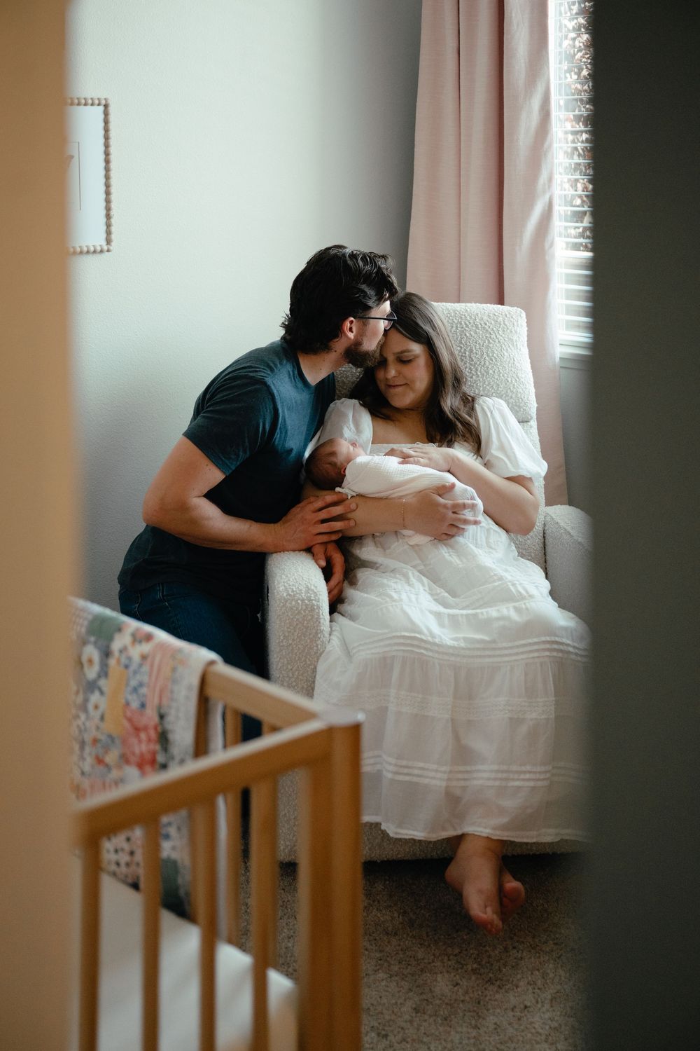 New parents sharing a tender moment with their newborn in a nursery, mother in white dress cradling baby while father kisses her head.