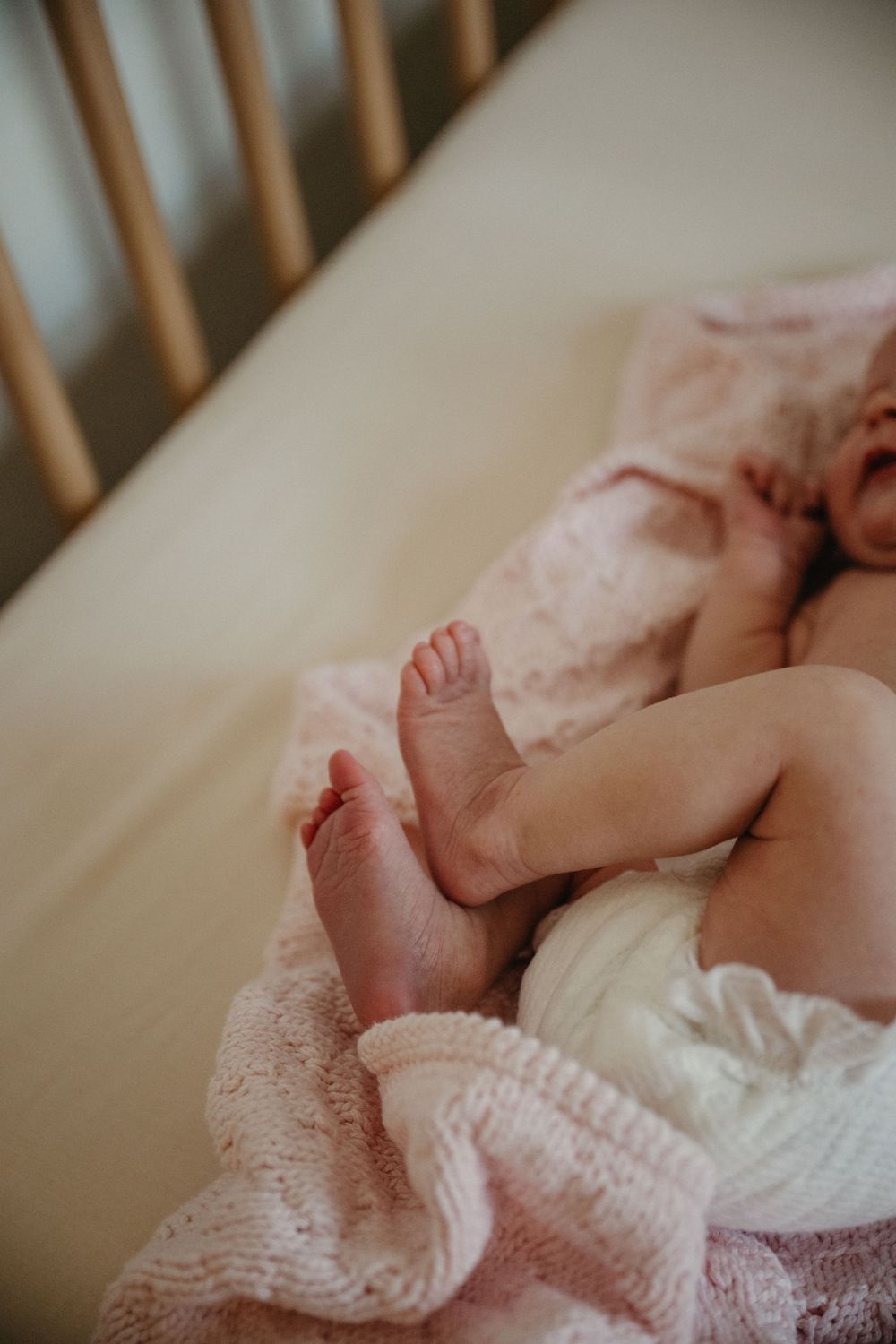 Newborn baby feet and legs resting on a soft pink knit blanket inside a wooden crib.