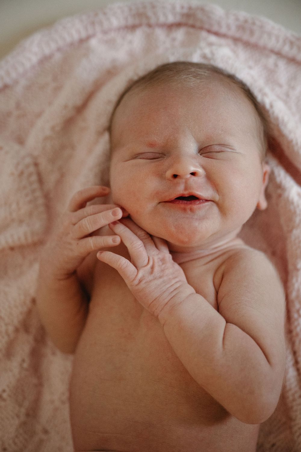 Peaceful newborn baby smiling in sleep, wrapped in a soft pink knit blanket, hands raised to cheek.