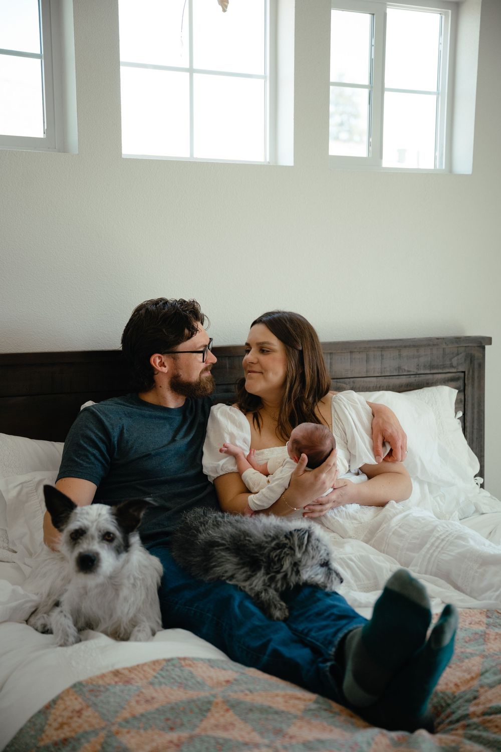 A couple sits on a bed with their newborn baby and two dogs, sharing a loving glance in a bright bedroom.