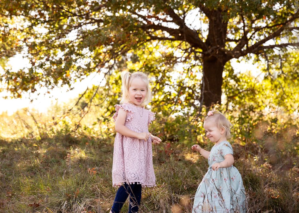 Two young children playing together in a sunny field under a large tree at golden hour.