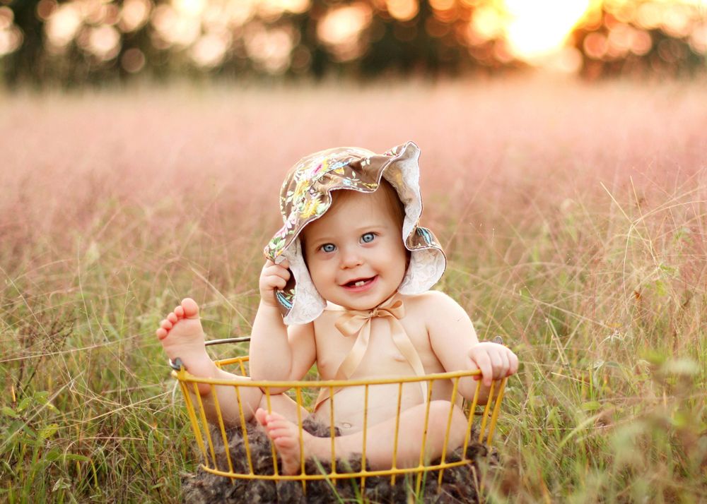 Baby sitting in vintage basket wearing bonnet during sunset in tall grass.
