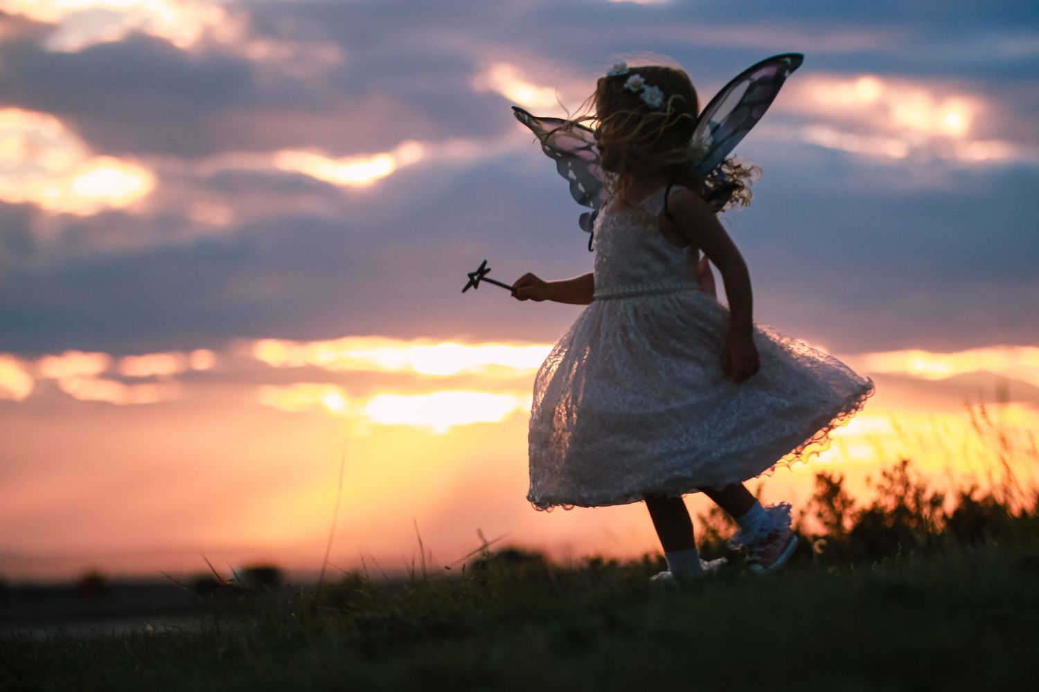 Young girl in white fairy costume with butterfly wings dancing in a field at golden sunset.