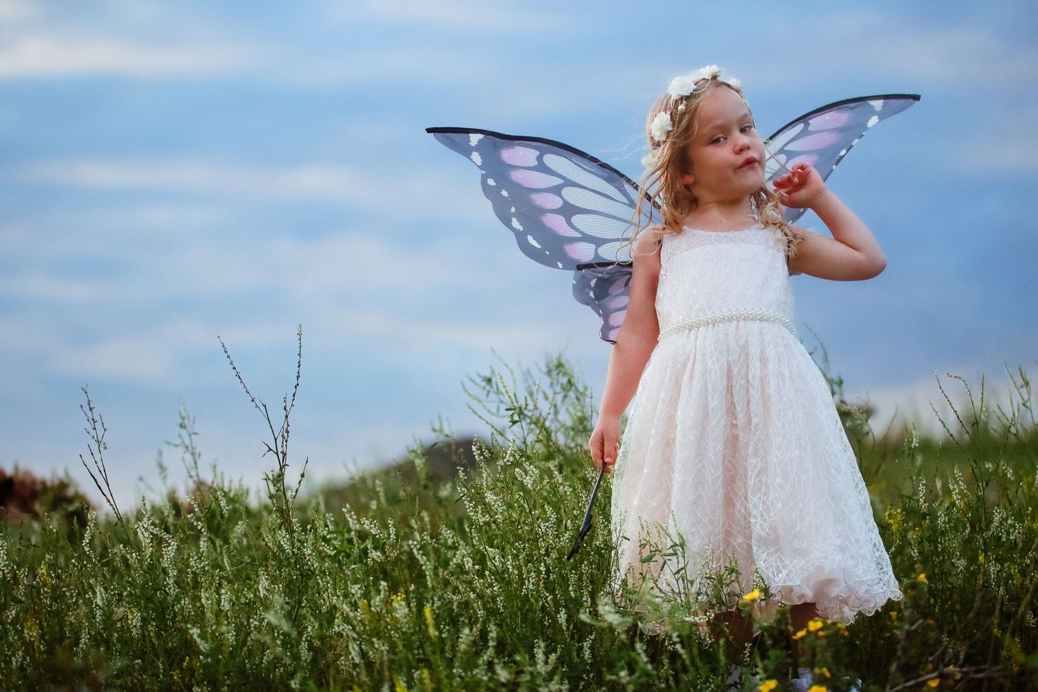 Little girl in white dress with purple butterfly wings standing in a green meadow under cloudy sky.