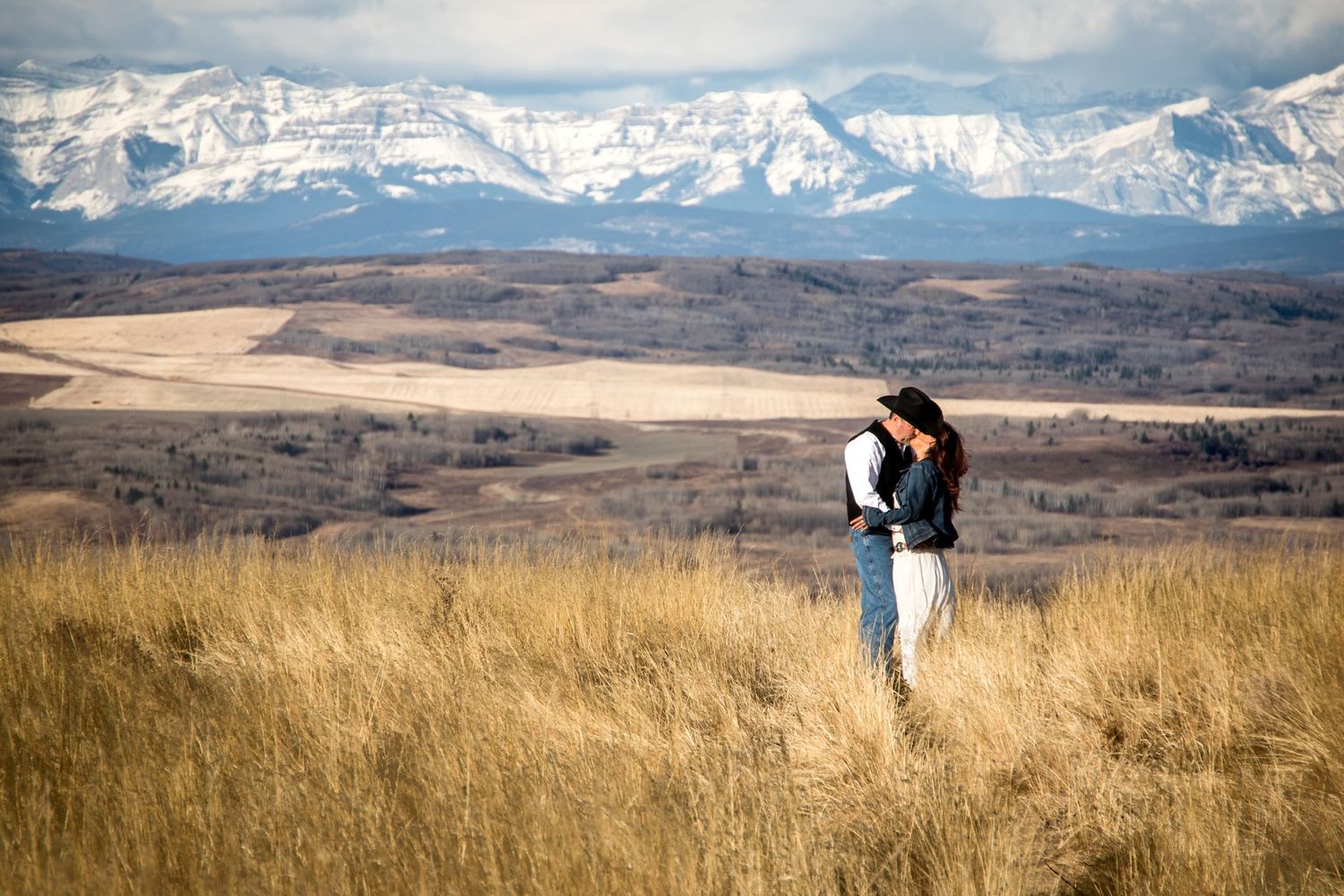 A woman stands in golden prairie grass with snow-capped Rocky Mountains in the background.