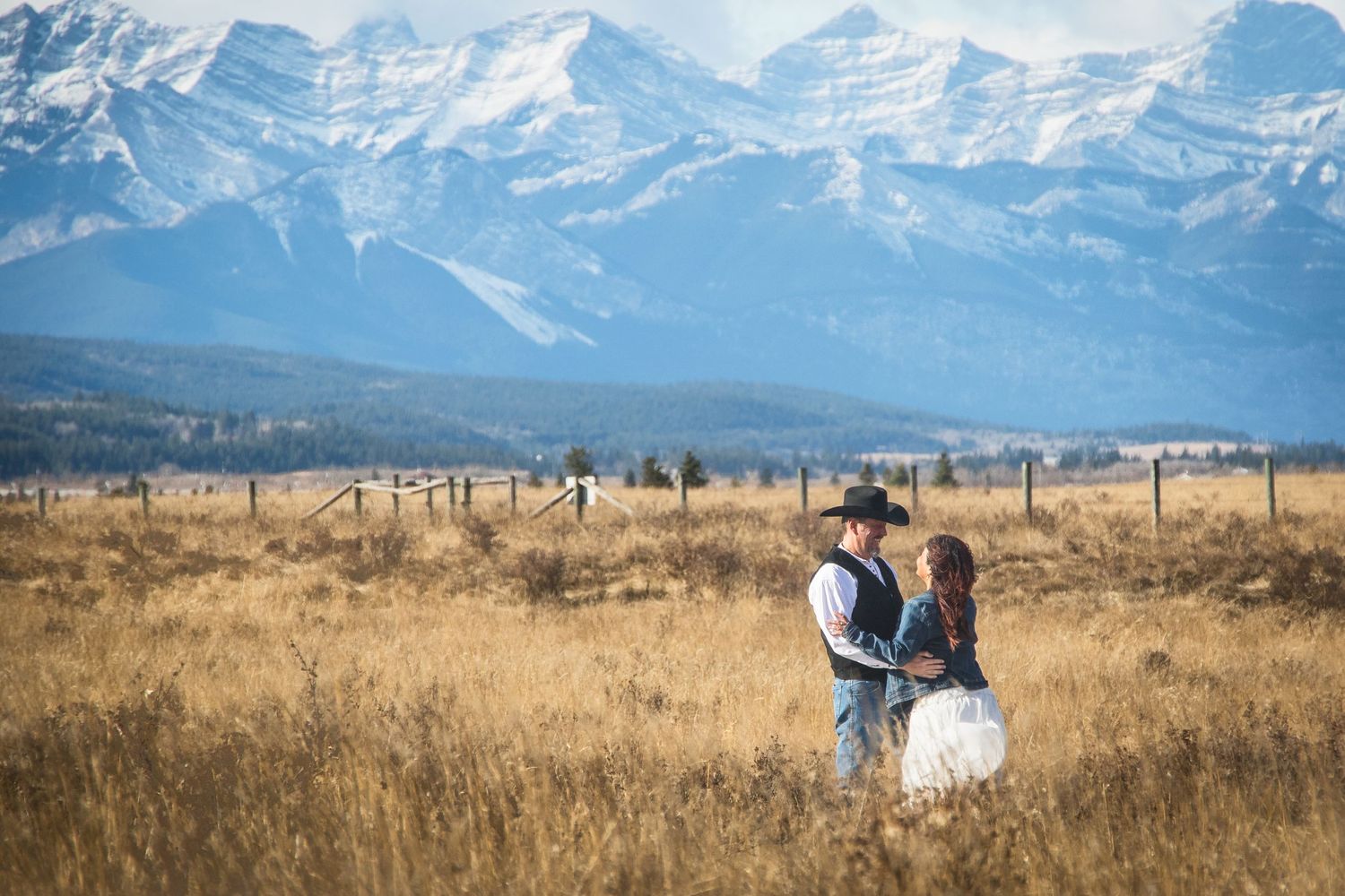 Couple embraces in golden prairie grass with stunning snow-capped Rocky Mountains in the background.