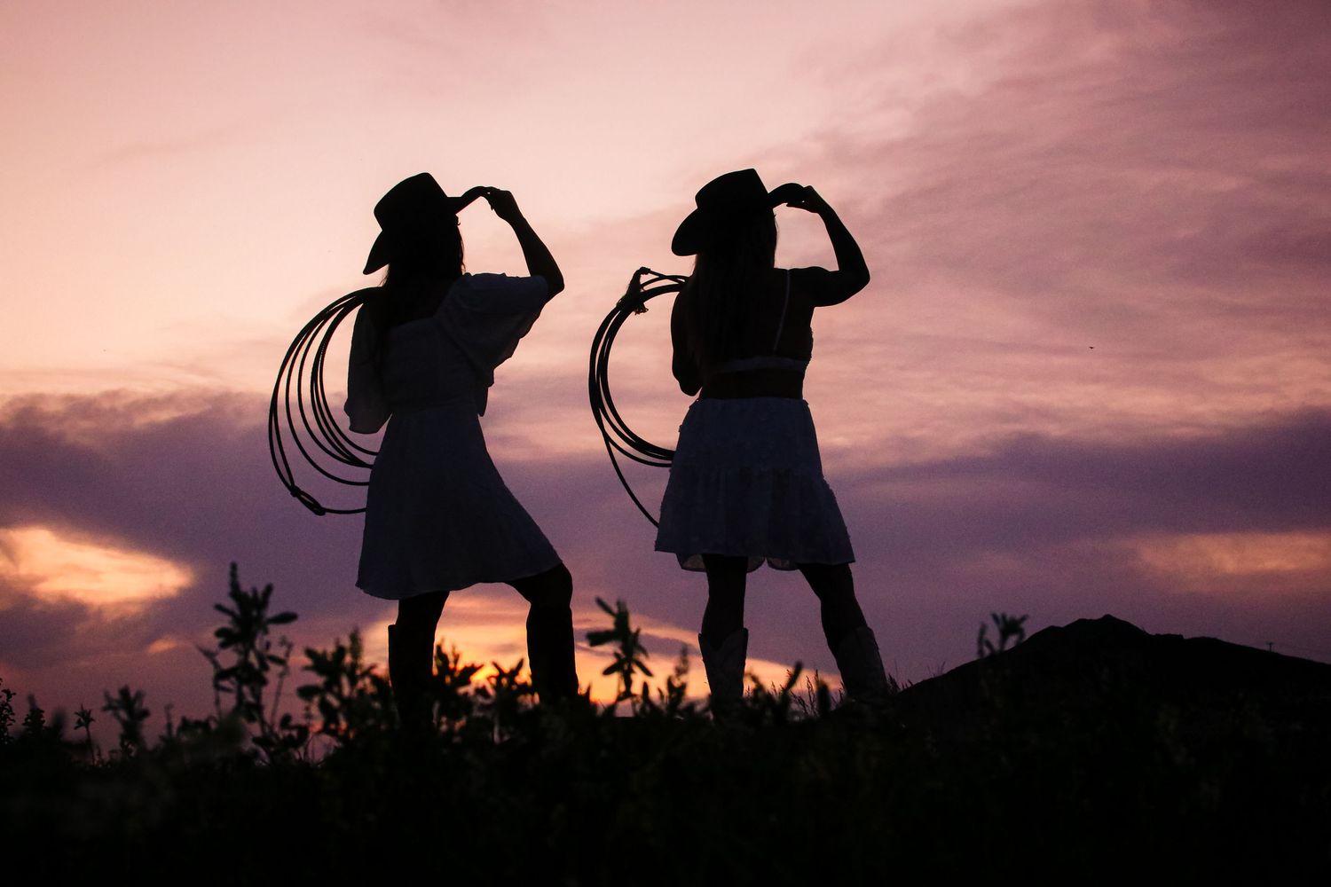 Silhouette of two cowgirls holding lassos and cowboy hats against a purple sunset sky.