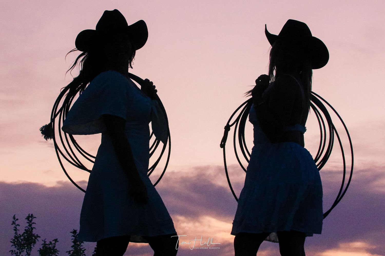 Two cowgirls in hats holding rope lassos posing against a dramatic pink and purple sunset.