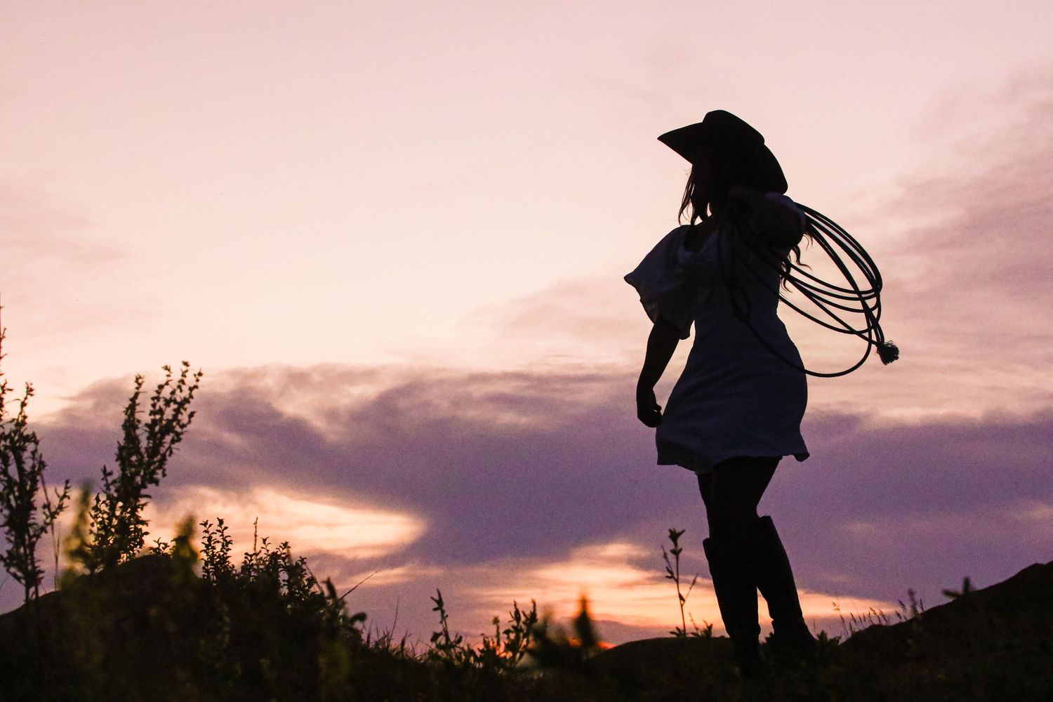 Silhouette of a cowgirl holding a lasso in a flowing dress against a purple cloudy sunset.