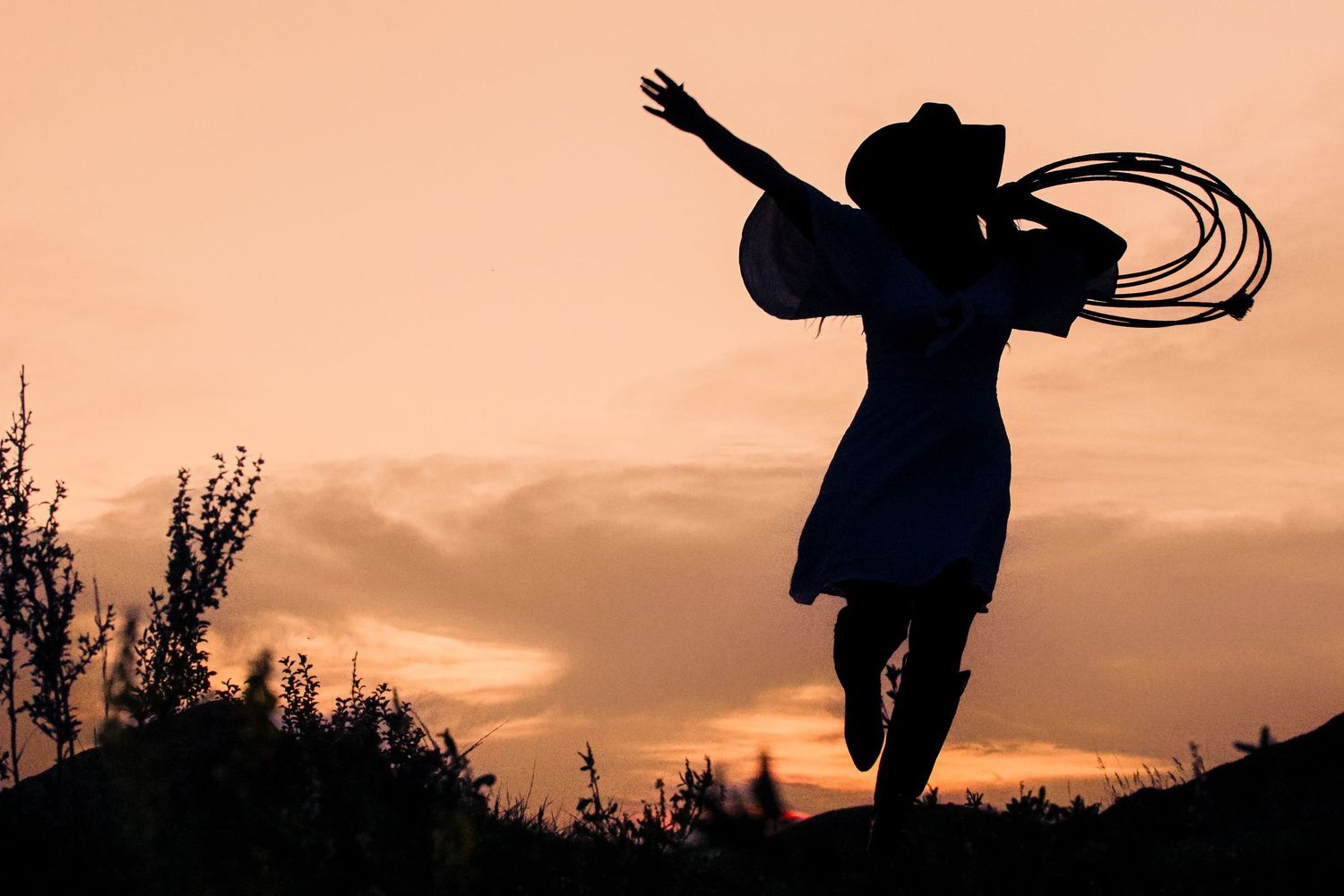 Silhouette of a cowgirl jumping joyfully with a lasso against a warm orange sunset sky.