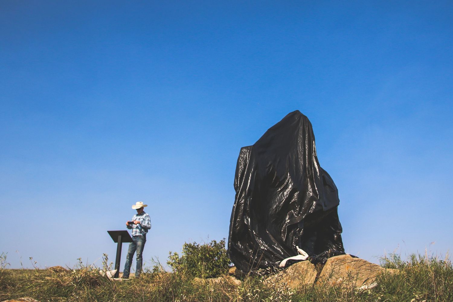 A person stands beside a large black draped sculpture under a vivid blue sky in an open field.