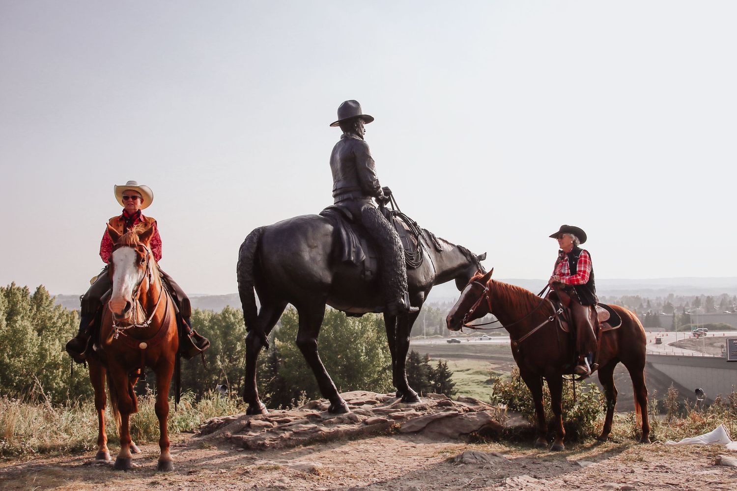 Two riders on horseback flank a bronze equestrian statue on a hilltop with a valley below.