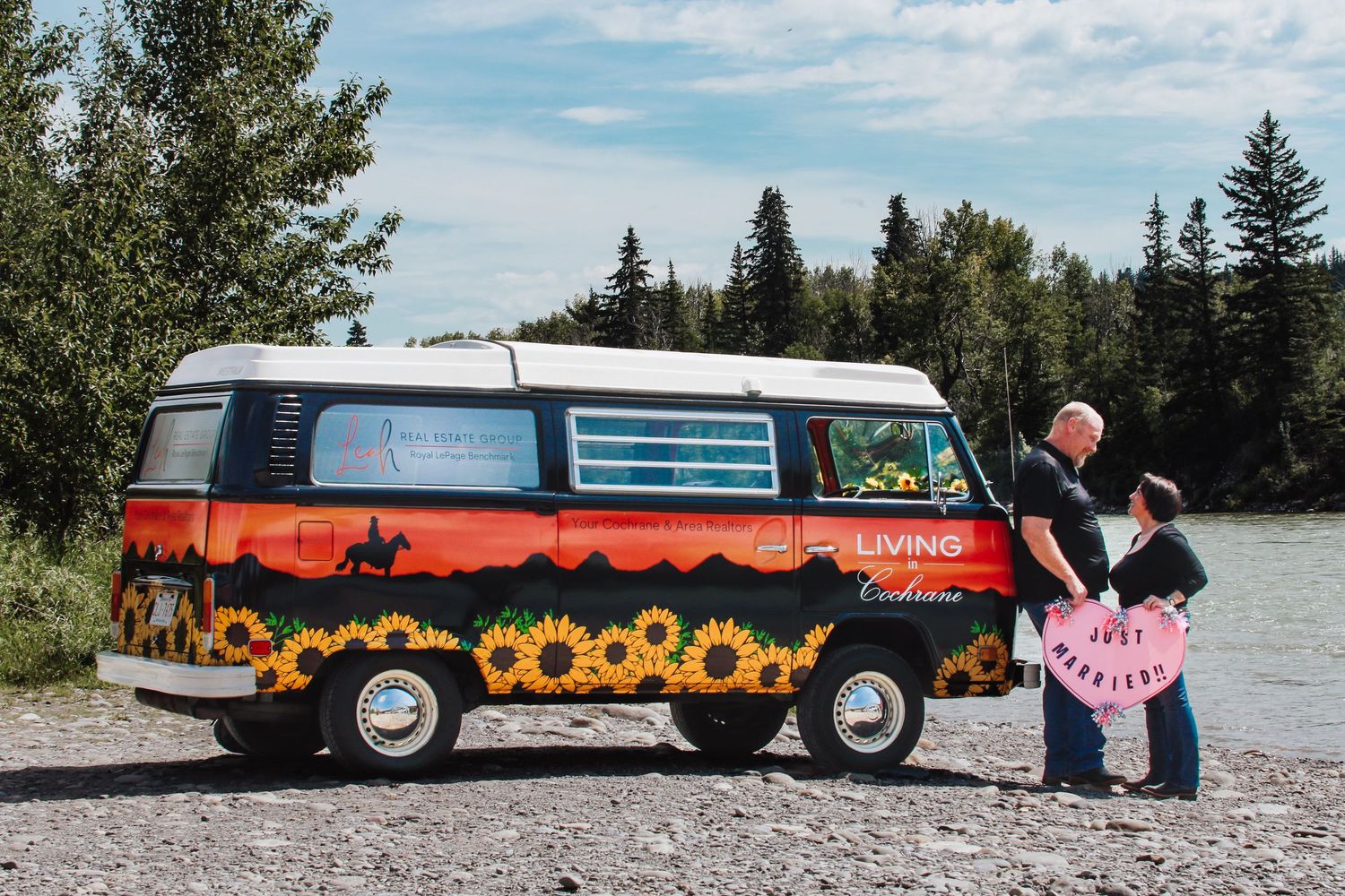 Colorful vintage VW bus with sunflower artwork parked by a river as a couple stands nearby.