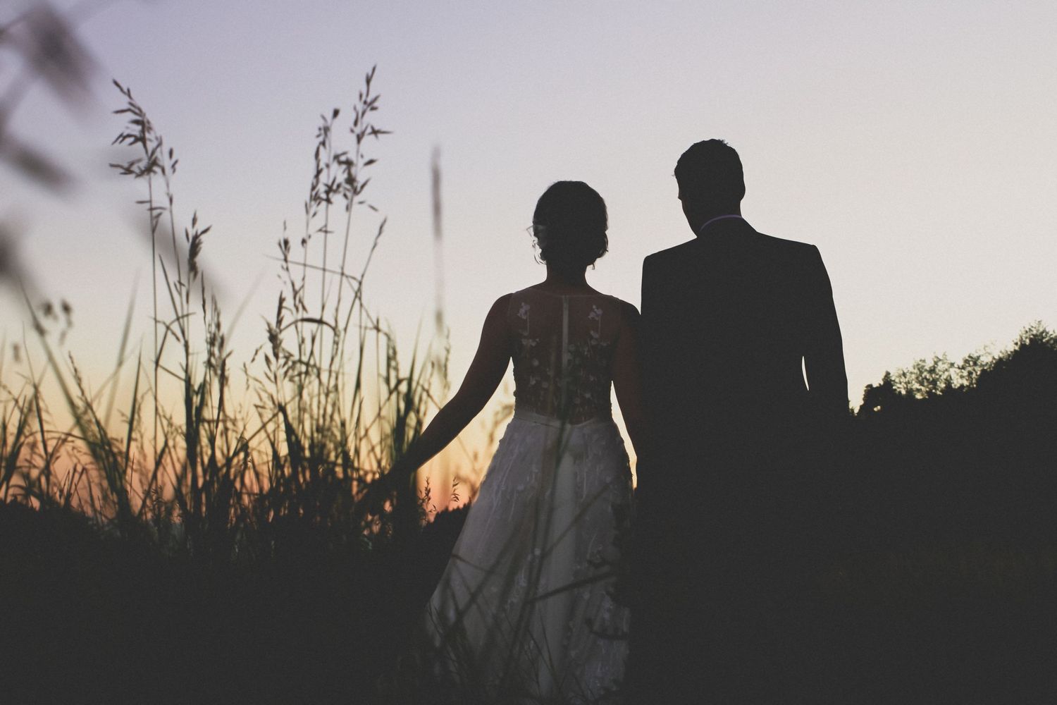Silhouette of a bride and groom holding hands in a field at sunset during their wedding.