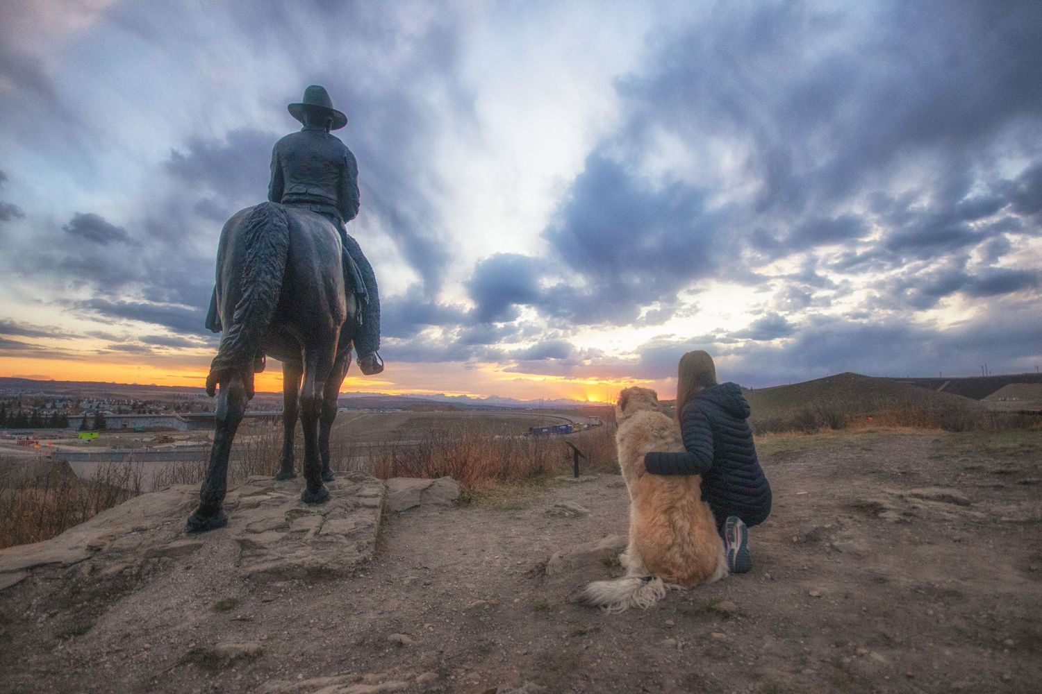A woman and her dog sit beside an equestrian statue at a scenic overlook during sunset.