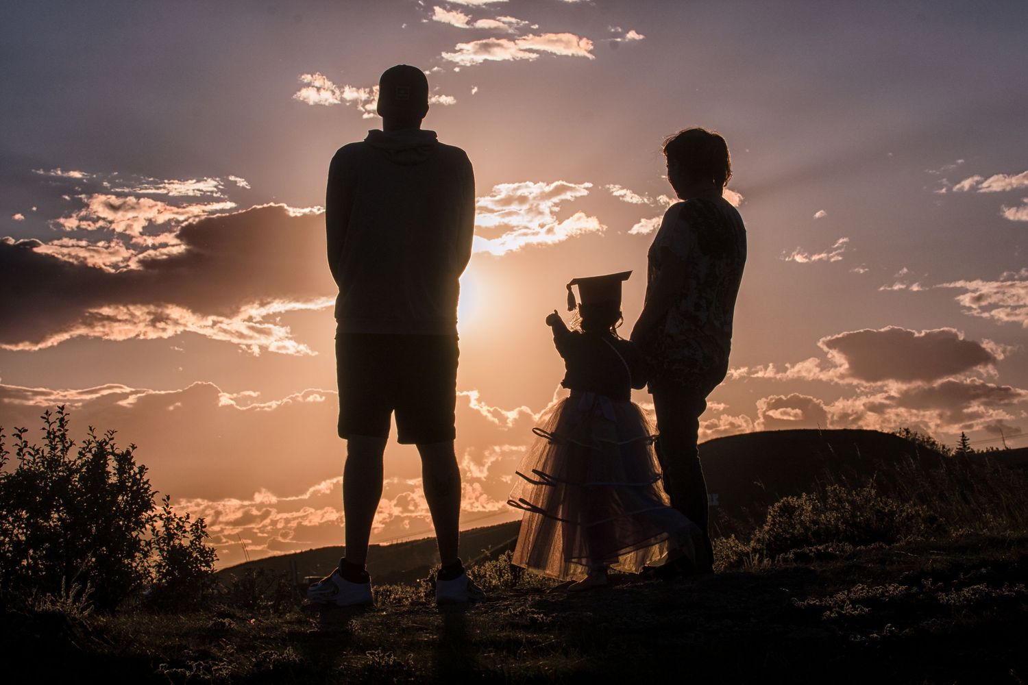 Silhouette of two adults and a child in a tutu watching a dramatic sunset from a hilltop.