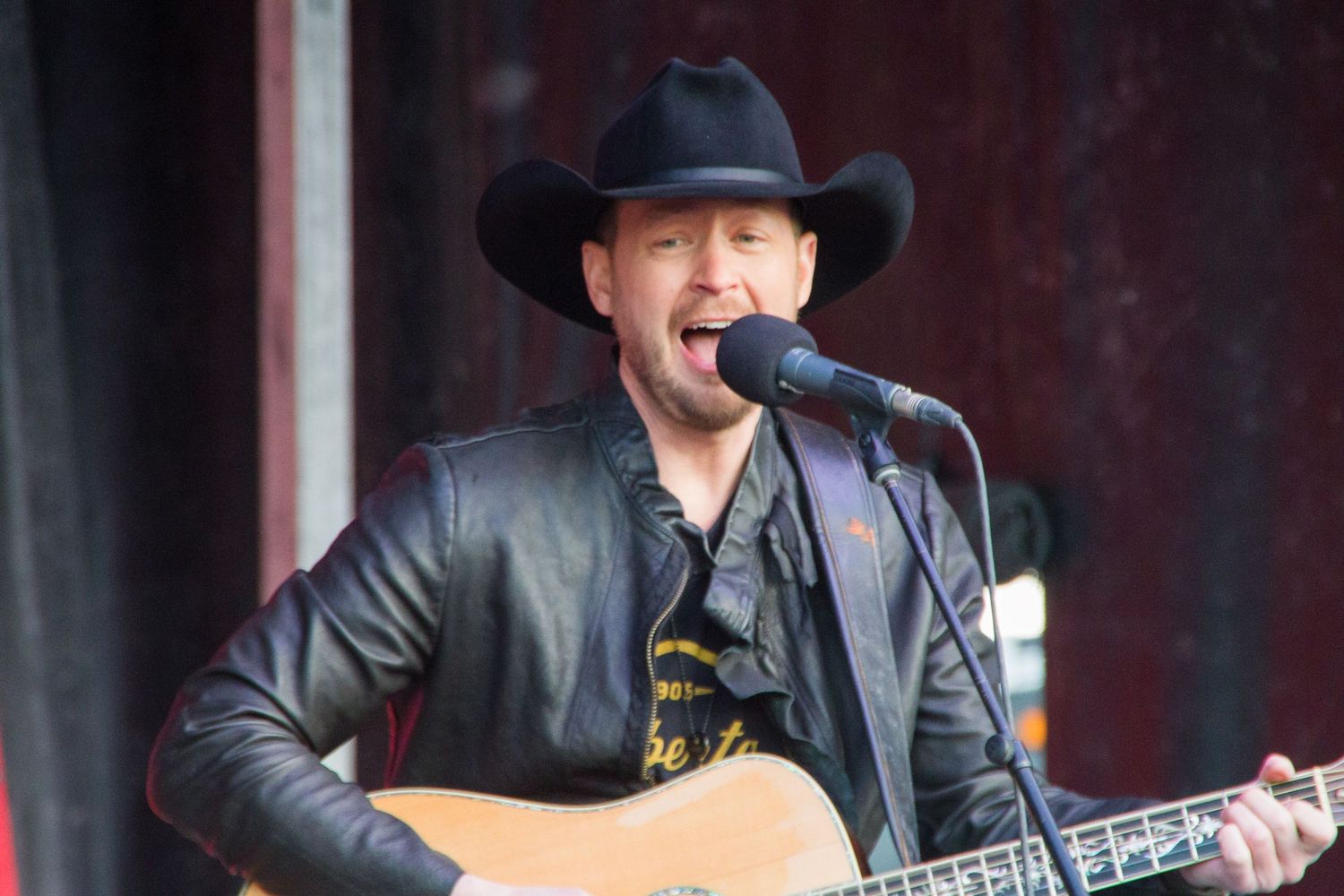 A country music performer in a black cowboy hat and jacket sings into a microphone while playing guitar.