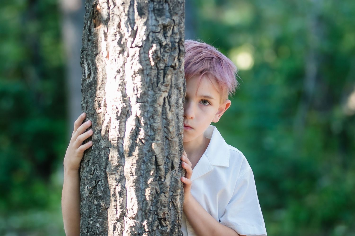 Young boy with pink hair peeks curiously around a tree trunk in a green forest setting.