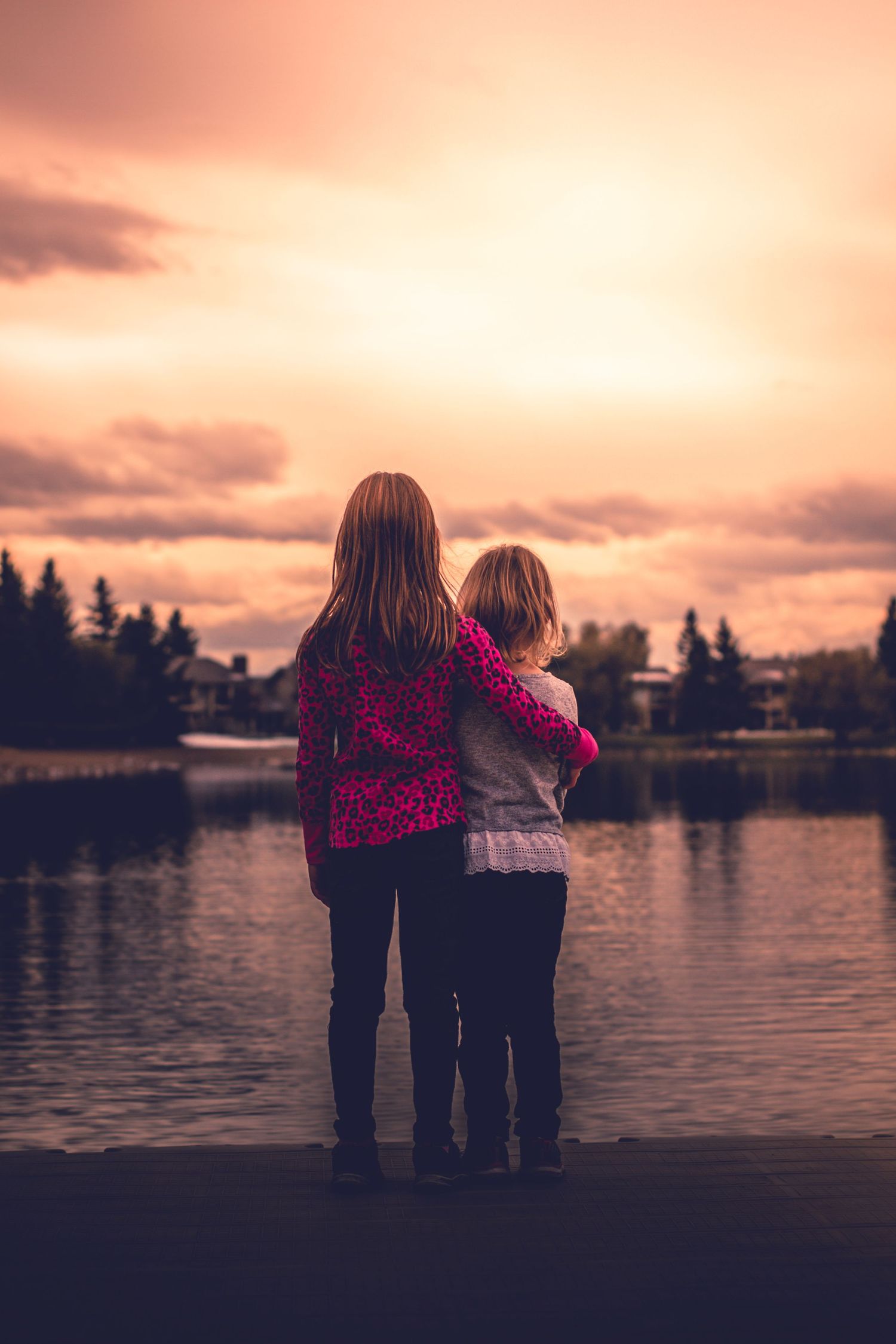 Two young girls embrace while watching a beautiful sunset over a calm lake surrounded by trees.