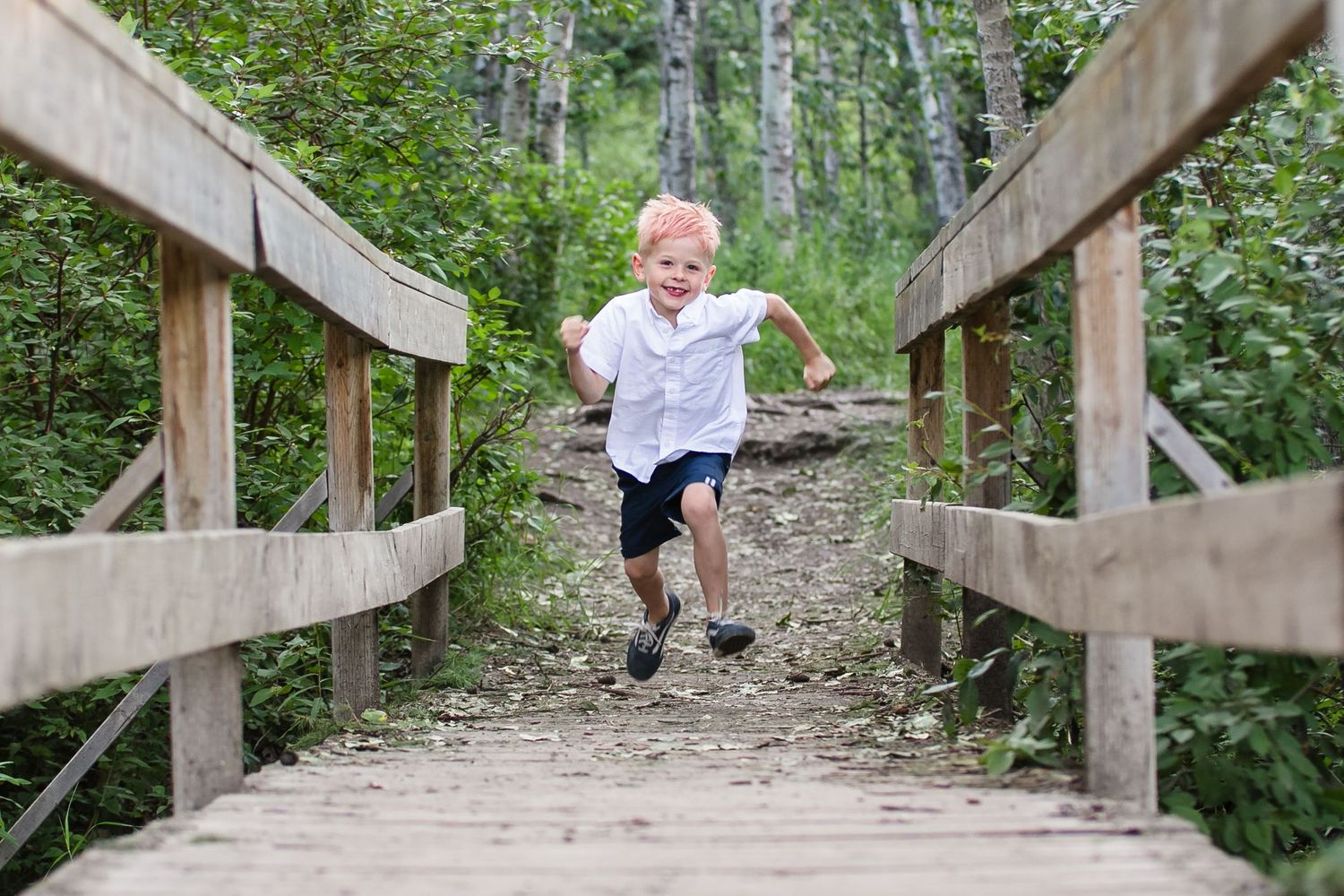Energetic young boy in a white shirt runs across a wooden bridge surrounded by lush green trees.