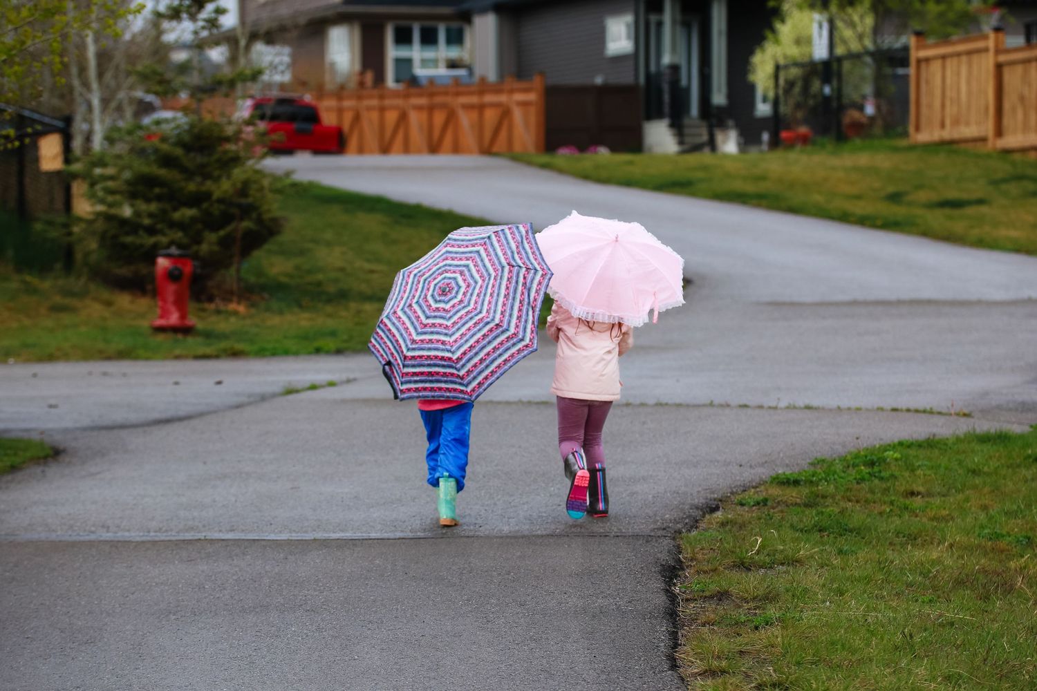 Two young children walk down a suburban sidewalk sharing colorful umbrellas on a rainy day.