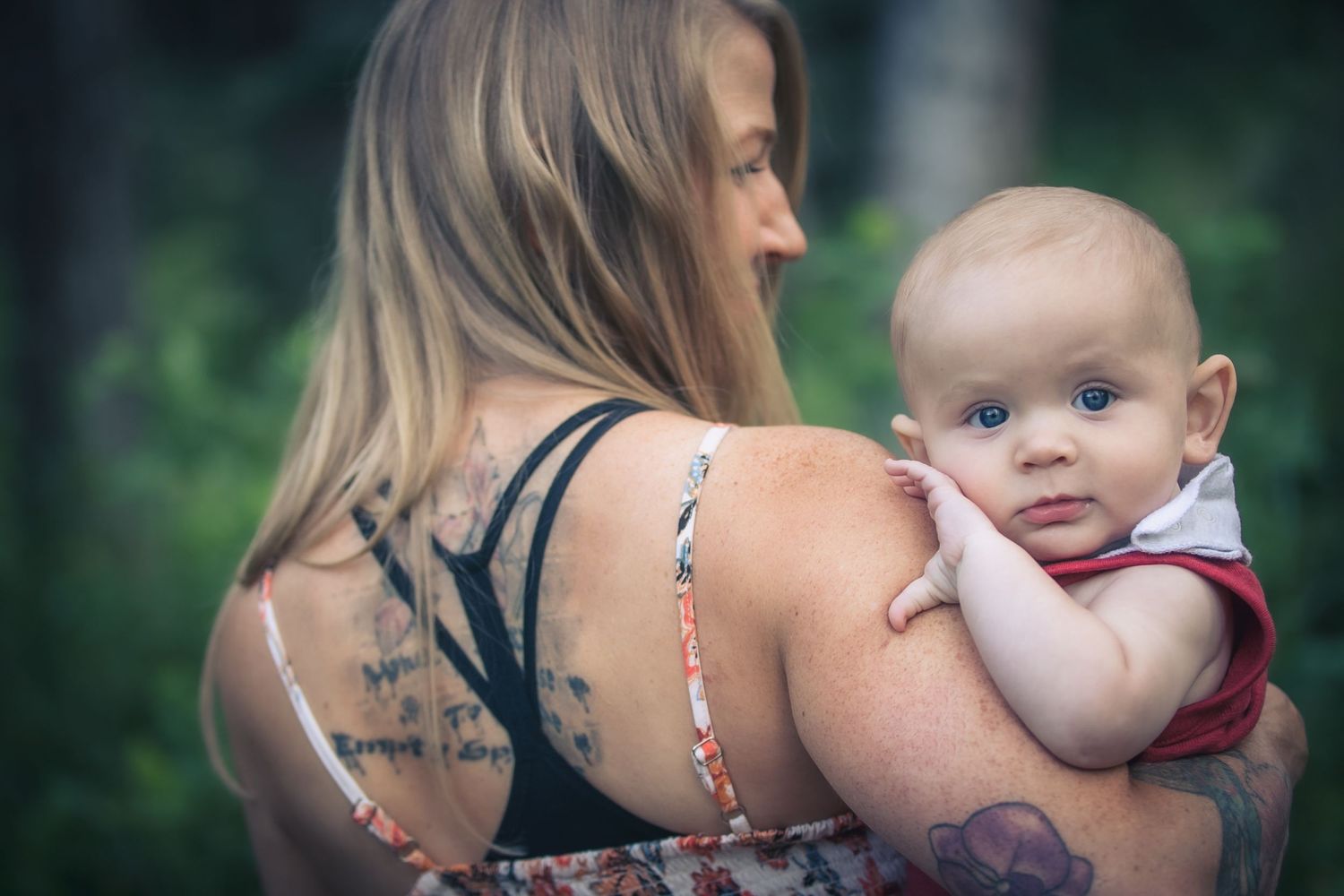 Tattooed mother holds her smiling baby over her shoulder in a soft bokeh green outdoor portrait.