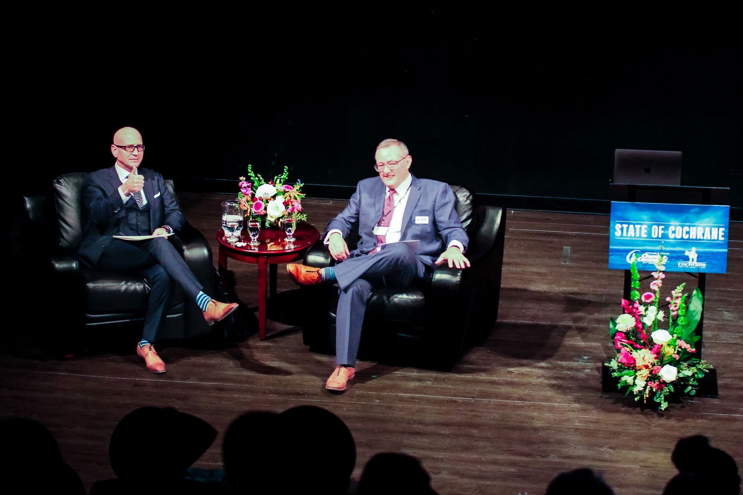 Two men in suits sit in black leather chairs on stage at a State of Cochrane event with floral arrangements.