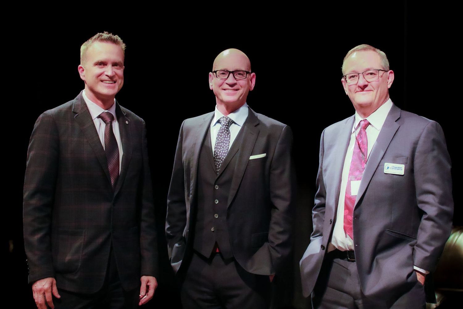 Three men in formal suits smile together on stage at a professional event in Cochrane.