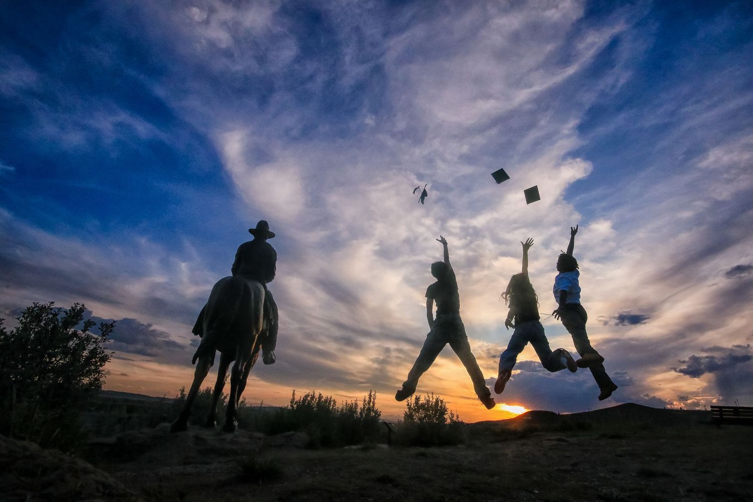 Silhouette of graduates jumping and tossing caps at sunset with a person on horseback nearby.