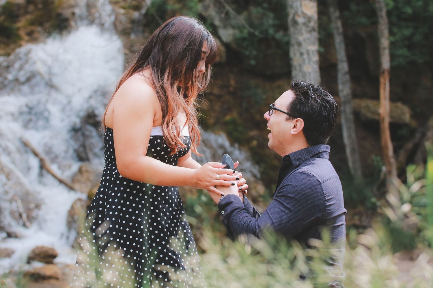 Man proposing to woman with engagement ring near a waterfall in the forest.