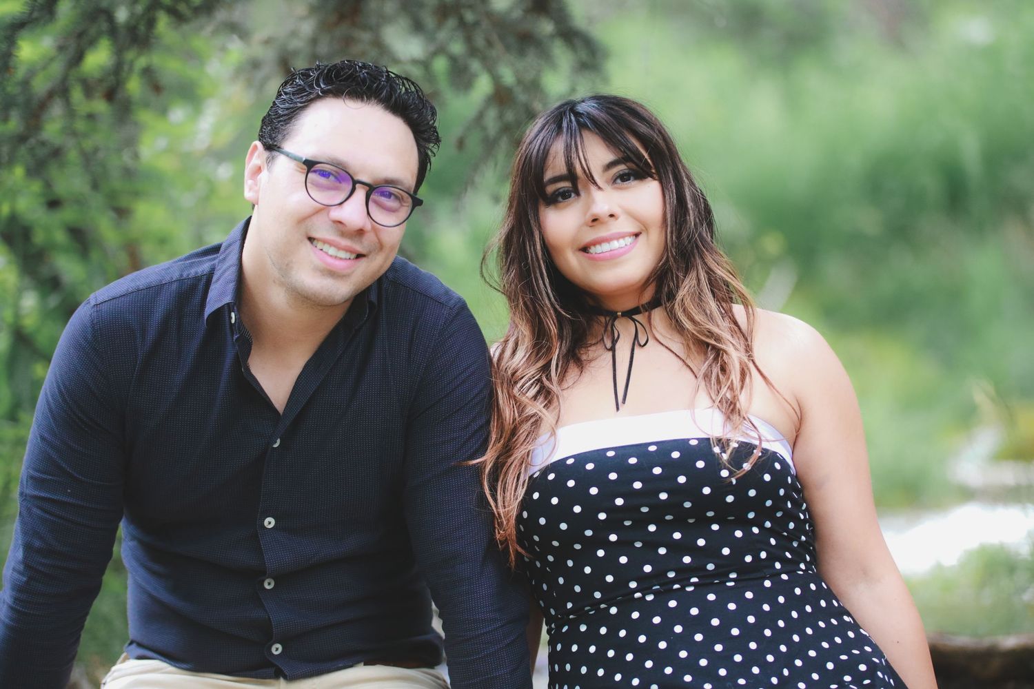 A smiling couple poses together outdoors in a lush green park setting.