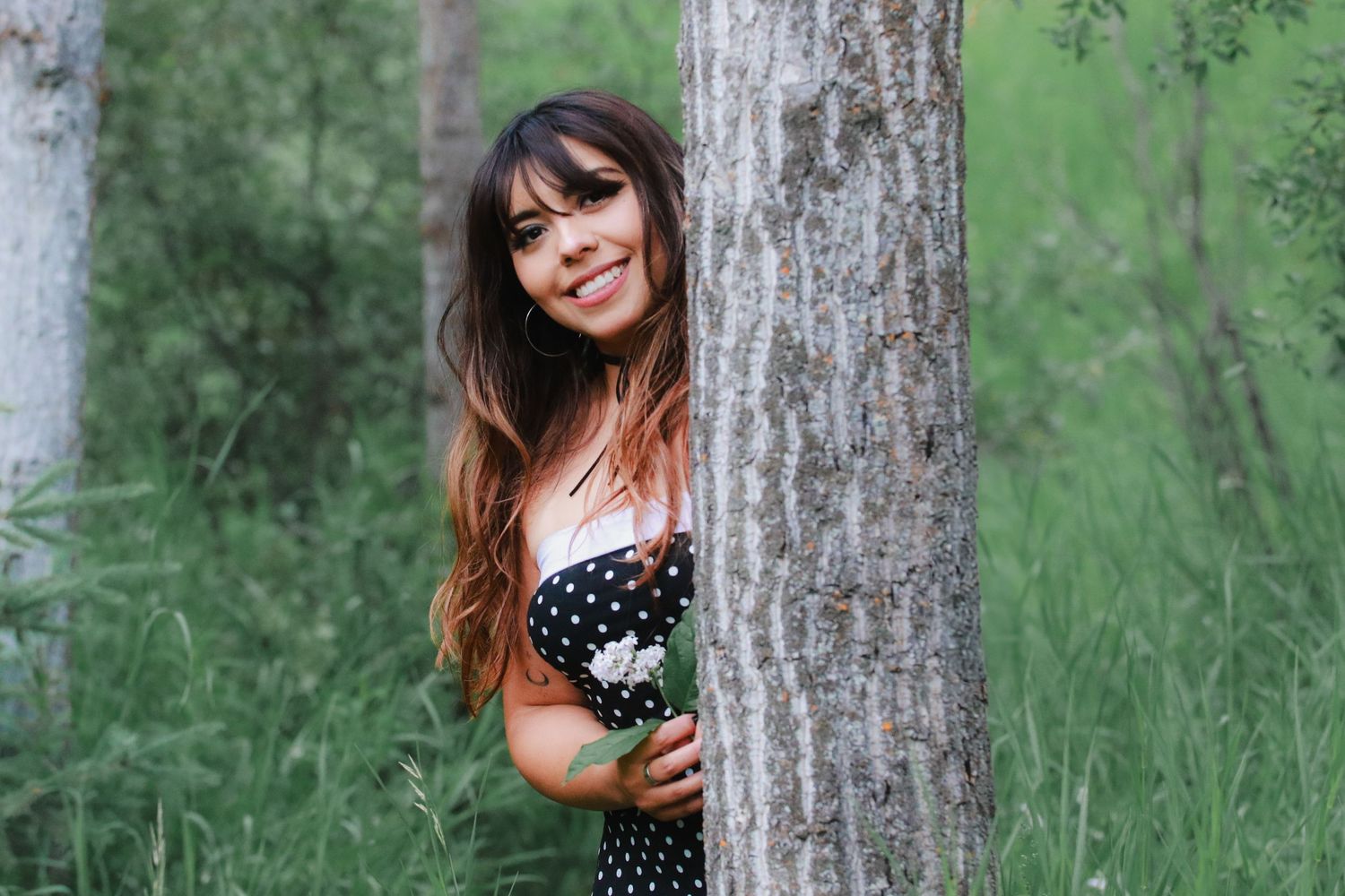 A young woman with long brown hair smiles while peeking from behind a tree in a forest.