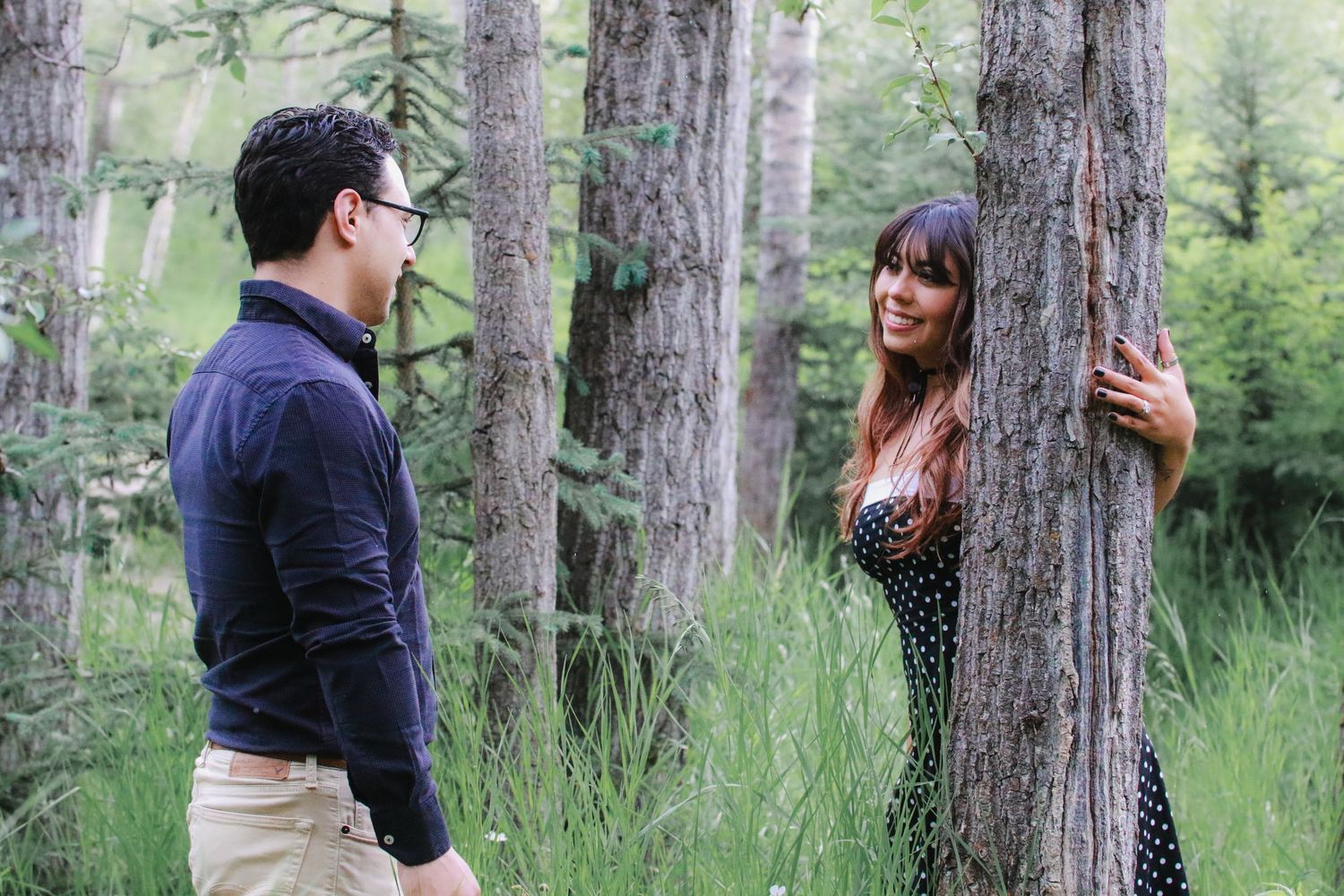 A man and woman interact playfully among tall trees in a green forest setting.
